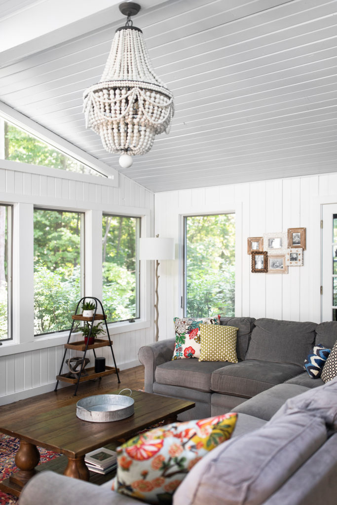 Grey sectional in white, open Hudson, OH Sunroom with beaded chandelier, wooden coffee table, and tired step ladder shelves Lindsey Putzier Design Studio