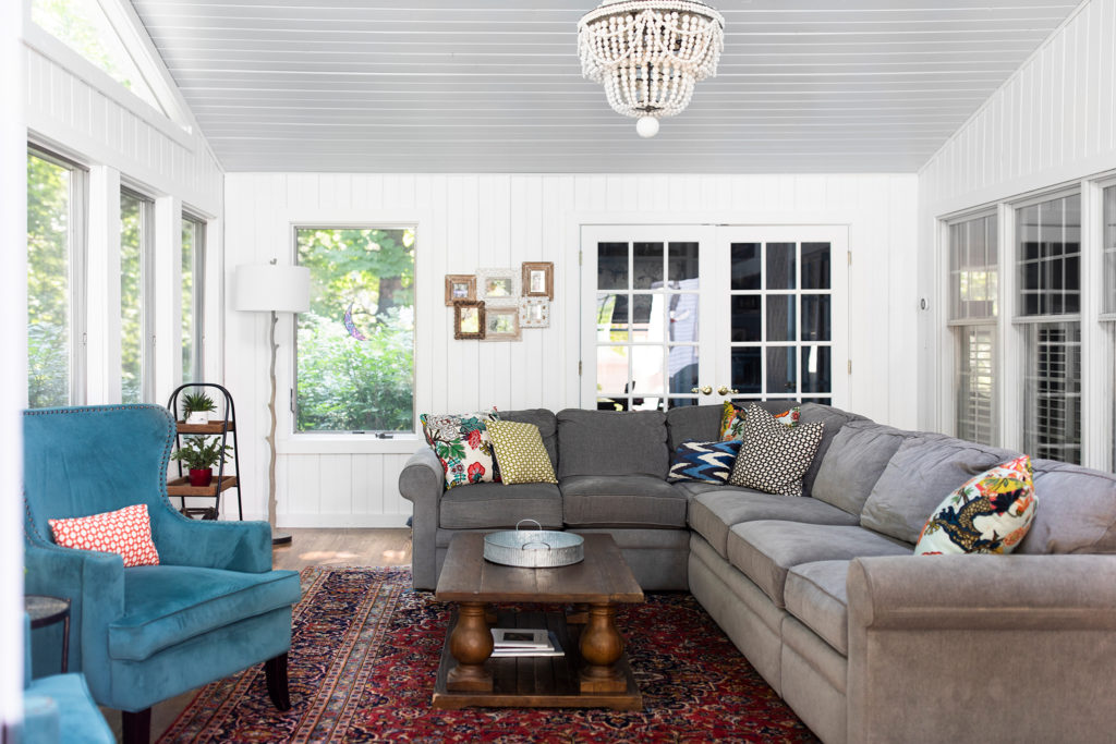 Gray sectional with accent pillows, teal lounge chairs, white beaded chandelier, red rug, and wooden coffee table Lindsey Putzier Design Studio